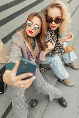 Гламурные молодые красивые девушки в модной одежде сидят на ступеньках,  пьют кофе и делают селфи на своих смартфонах | Премиум Фото